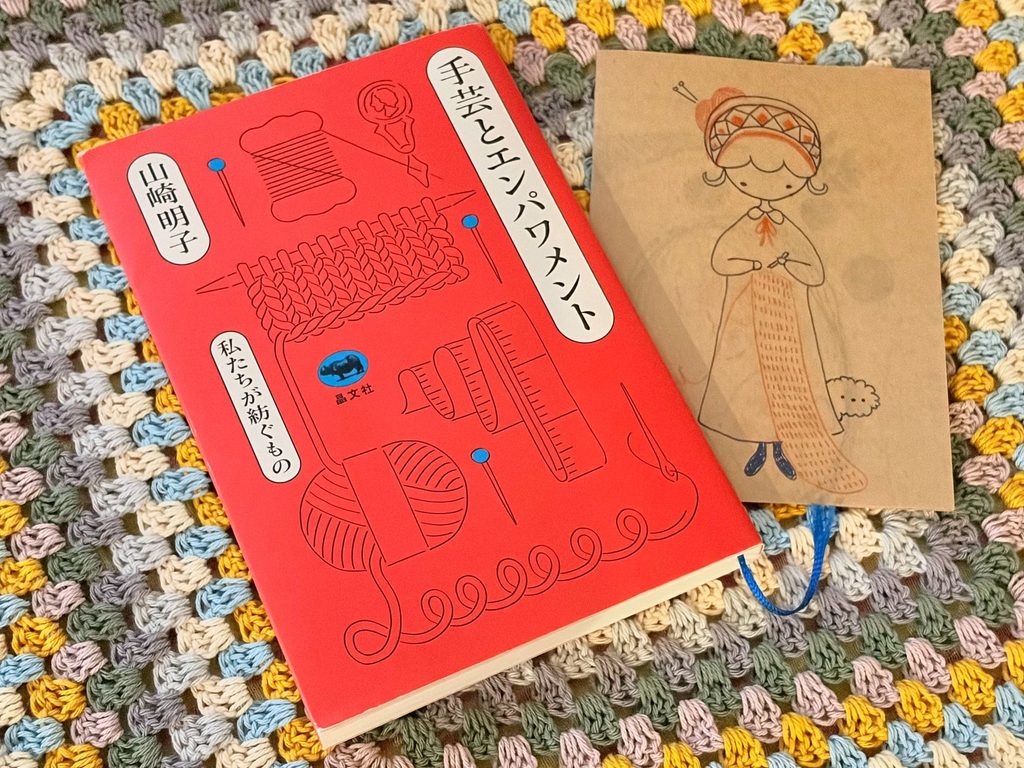 編み物の上に置いた、山崎明子さんの著書『手芸とエンパワメント』と、女性の刺しゅうが入ったブックカバー